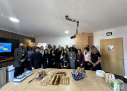 A large group of diverse professionals gathered for a celebration in an office meeting room. The team stands behind a wooden table filled with cakes, pastries, and snacks. A presentation screen is visible in the background, and everyone is dressed in business attire wearing ID badges.