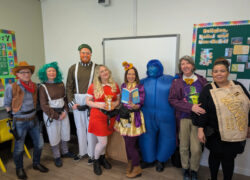 A group of people dressed in various costumes standing in front of a whiteboard in a classroom. Costumes include what appear to be characters from children's literature, including someone in a blue full-body costume, another in a purple coat, someone dressed as a cowboy, and others in themed outfits. A colorful educational bulletin board is visible in the background.