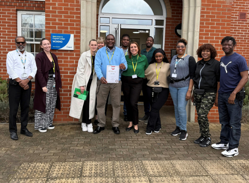 Group of diverse professionals standing in front of a brick building entrance. A person in the centre is holding a accreditation certificate.