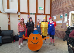 A group of people wearing costumes in what appears to be a community hall. The costumes include Where's Waldo (striped shirt and hat), Wednesday Adams (black dress and braids), a person in a minion costume, someone dressed as a yellow cheese or LEGO block, and Mario (in red overalls). In the foreground is a person in an orange Mr. Men/Little Miss character costume. The room has brick walls, a notice board, and an emergency exit sign