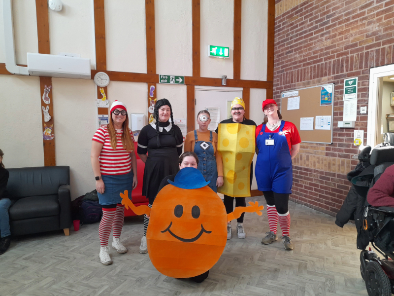 A group of people wearing costumes in what appears to be a community hall. The costumes include Where's Waldo (striped shirt and hat), Wednesday Adams (black dress and braids), a person in a minion costume, someone dressed as a yellow cheese or LEGO block, and Mario (in red overalls). In the foreground is a person in an orange Mr. Men/Little Miss character costume. The room has brick walls, a notice board, and an emergency exit sign