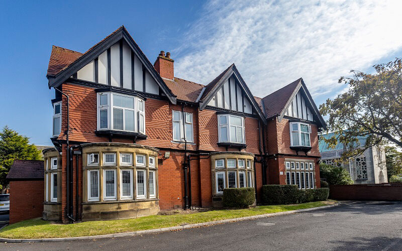 A stately Tudor-style building with distinctive architectural features. The structure has red brick walls with decorative dark timber framing on the upper levels and prominent gabled roofs. There are multiple bay windows with white frames, and the building features a large chimney