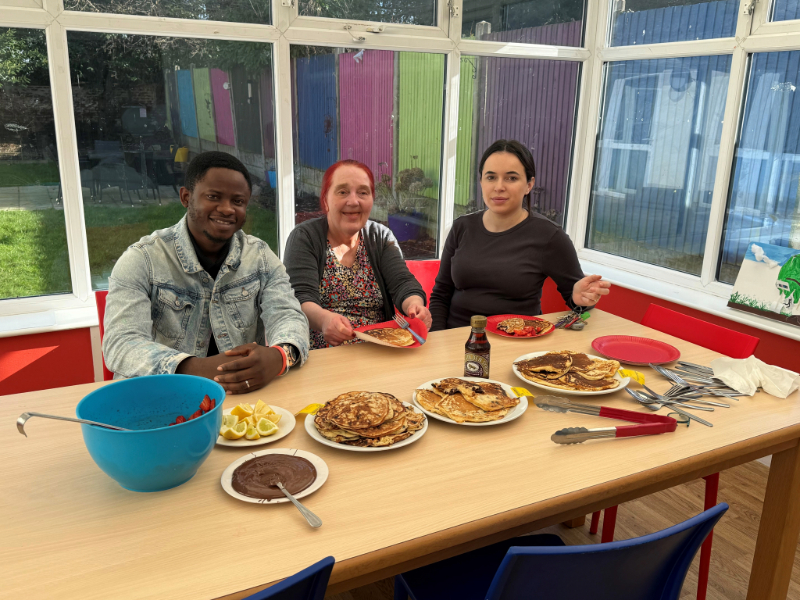 Three people sitting at a table with pancakes, smiling - a man in denim jacket, a woman with red hair, and another woman in a black top