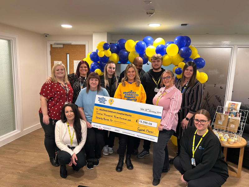 A group of staff standing together indoors, holding a large presentation cheque for Sheffield Children’s Hospital Charity. Behind them is a decorative arch made of blue and yellow balloons, and a table of branded cupcakes is visible to the side.