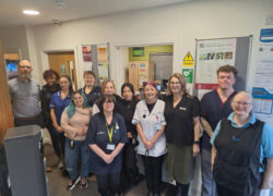 A group of healthcare staff stand together in a clinical reception area, wearing a mix of uniforms and work attire. They are positioned in front of noticeboards, posters and safety signs, with a small office window behind them displaying paperwork and supplies. The team is gathered closely for a group photo.