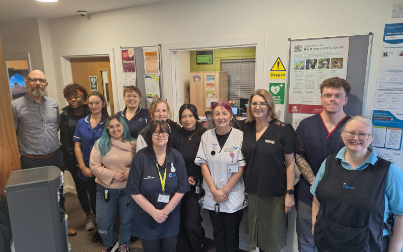 A group of healthcare staff stand together in a clinical reception area, wearing a mix of uniforms and work attire. They are positioned in front of noticeboards, posters and safety signs, with a small office window behind them displaying paperwork and supplies. The team is gathered closely for a group photo.