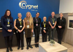 Seven staff members standing in a row inside Cygnet Hospital Derby reception. They are wearing work attire with lanyards and ID badges. Behind them is a blue wall with the Cygnet Health Care logo in large silver letters. In front, there is a light-coloured table with a small potted plant.