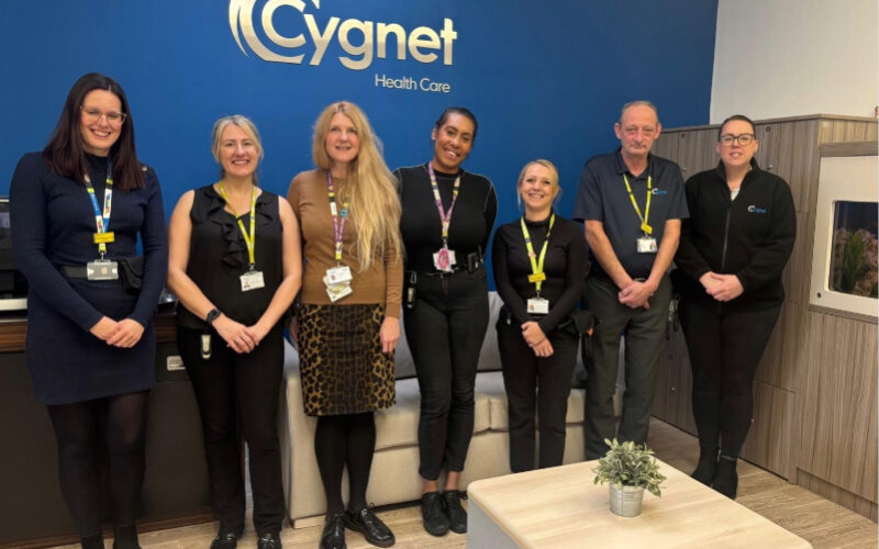Seven staff members standing in a row inside Cygnet Hospital Derby reception. They are wearing work attire with lanyards and ID badges. Behind them is a blue wall with the Cygnet Health Care logo in large silver letters. In front, there is a light-coloured table with a small potted plant.