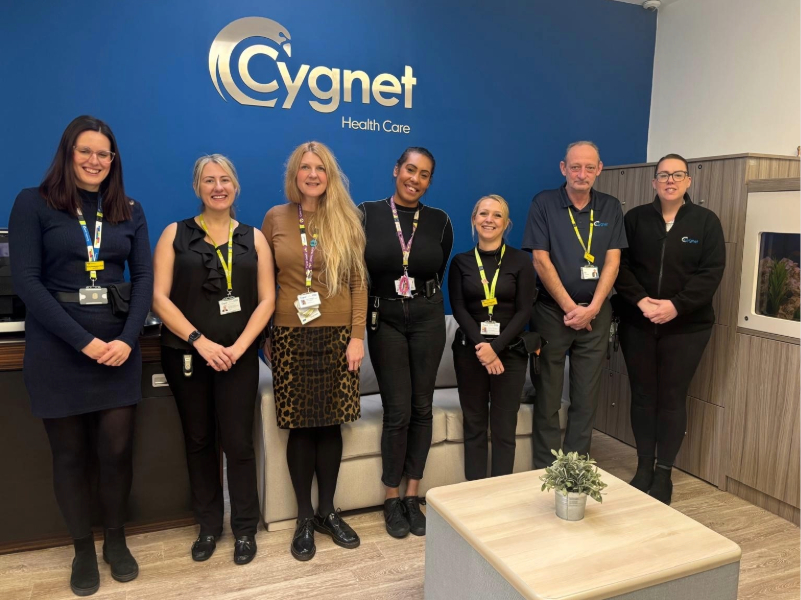 Seven staff members standing in a row inside Cygnet Hospital Derby reception. They are wearing work attire with lanyards and ID badges. Behind them is a blue wall with the Cygnet Health Care logo in large silver letters. In front, there is a light-coloured table with a small potted plant.