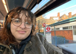 Person sitting on a bus wearing headphones and a green jacket, with a view through the window showing a London bus stop sign and timetable.