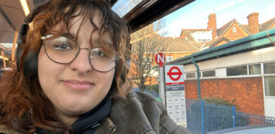 Person sitting on a bus wearing headphones and a green jacket, with a view through the window showing a London bus stop sign and timetable.