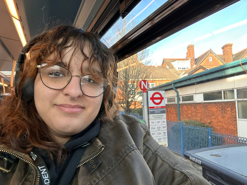Person sitting on a bus wearing headphones and a green jacket, with a view through the window showing a London bus stop sign and timetable.