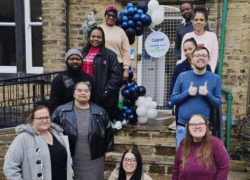 A group of people standing on stone steps outside a brick building decorated with a balloon arch in dark blue and white. A circular sign on the door reads “Cygnet Langdale House Opening.” The setting is an entrance area with a metal gate behind the balloons and a green pipe along the wall.