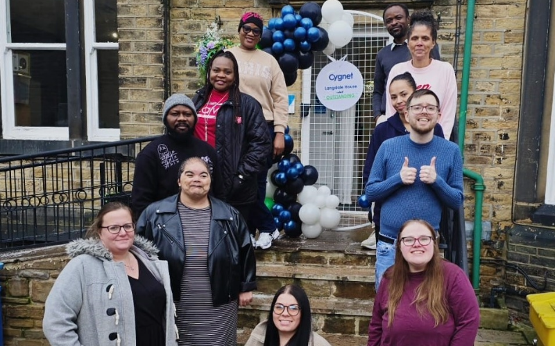 A group of people standing on stone steps outside a brick building decorated with a balloon arch in dark blue and white. A circular sign on the door reads “Cygnet Langdale House Opening.” The setting is an entrance area with a metal gate behind the balloons and a green pipe along the wall.