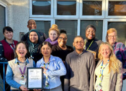 Group of staff standing outside a building with large windows, holding a framed Triangle of Care certificate. Several people are wearing lanyards and work attire, and the group is gathered closely together in bright daylight.