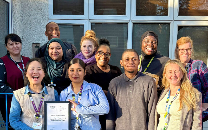 Group of staff standing outside a building with large windows, holding a framed Triangle of Care certificate. Several people are wearing lanyards and work attire, and the group is gathered closely together in bright daylight.