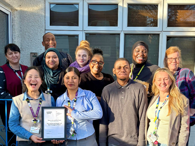 Group of staff standing outside a building with large windows, holding a framed Triangle of Care certificate. Several people are wearing lanyards and work attire, and the group is gathered closely together in bright daylight.