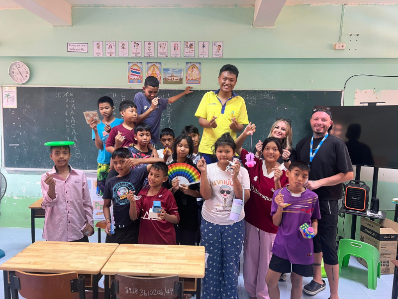A large group of Thai children and Ben and Holly pose together in a classroom, holding colourful toys, craft items and supplies in front of a chalkboard decorated with drawings and posters.