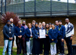 The Tyler Ward team is wearing uniforms and ID lanyards as they gather outside to celebrate Cygnet Hospital Maidstone receiving QNPICU accreditation from the Royal College of Psychiatrists. One person in the centre holds a framed accreditation certificate.