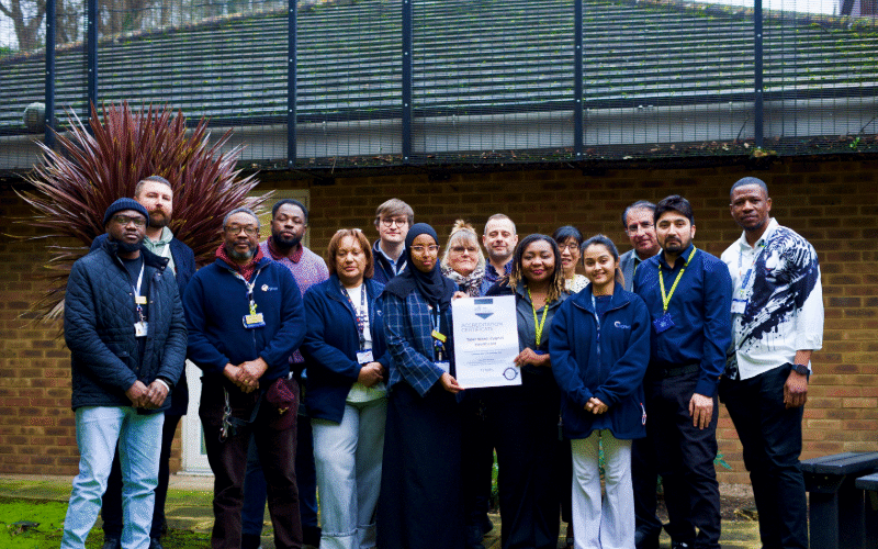 The Tyler Ward team is wearing uniforms and ID lanyards as they gather outside to celebrate Cygnet Hospital Maidstone receiving QNPICU accreditation from the Royal College of Psychiatrists. One person in the centre holds a framed accreditation certificate.