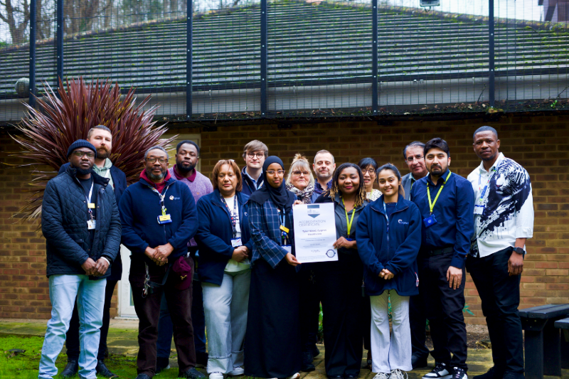 The Tyler Ward team is wearing uniforms and ID lanyards as they gather outside to celebrate Cygnet Hospital Maidstone receiving QNPICU accreditation from the Royal College of Psychiatrists. One person in the centre holds a framed accreditation certificate. 