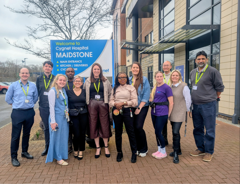 A group of staff members stand together outside Cygnet Hospital Maidstone, posing in front of a large blue sign that reads “Welcome to Cygnet Hospital Maidstone.” The team is gathered on a paved area beside the hospital building, smiling and wearing their ID lanyards. They are celebrating the hospital’s upgraded CQC rating of ‘Good’.