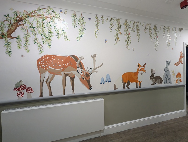 A bright, colourful woodland mural featuring illustrated animals such as a deer, fox and rabbits, with hanging greenery across the top of the wall in a corridor at Conifers