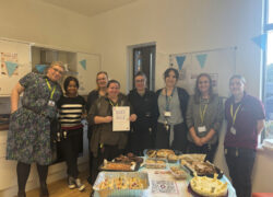 Members of the Cygnet Acer Clinic team standing together behind a table filled with homemade cakes, brownies and pastries. One person in the middle is holding a “Bake Sale” sign.