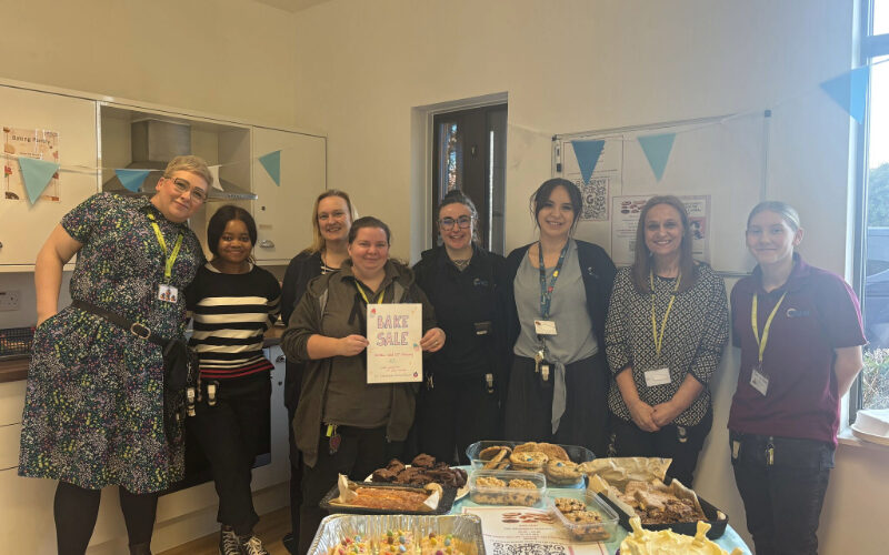 Members of the Cygnet Acer Clinic team standing together behind a table filled with homemade cakes, brownies and pastries. One person in the middle is holding a “Bake Sale” sign.