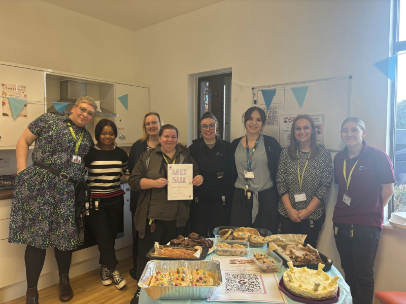 Members of the Cygnet Acer Clinic team standing together behind a table filled with homemade cakes, brownies and pastries. One person in the middle is holding a “Bake Sale” sign.