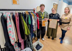 Three members of the Cygnet Hospital Kewstoke team smiling beside a clothing rail inside The Shop Upstairs. The rail displays a selection of donated clothing.