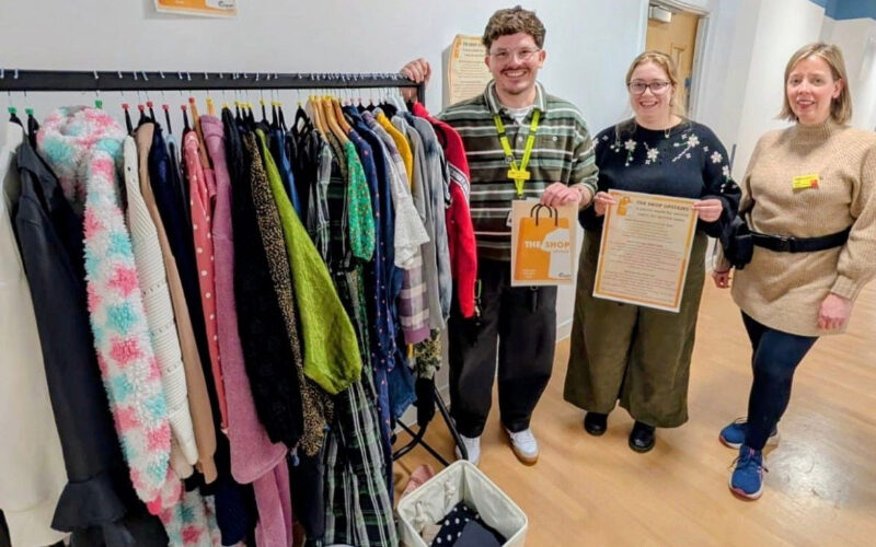 Three members of the Cygnet Hospital Kewstoke team smiling beside a clothing rail inside The Shop Upstairs. The rail displays a selection of donated clothing.