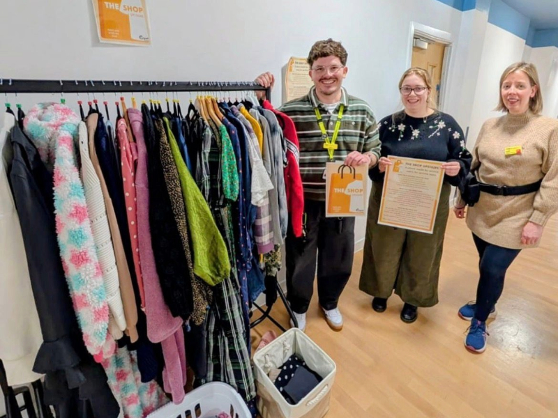 Three members of the Cygnet Hospital Kewstoke team smiling beside a clothing rail inside The Shop Upstairs. The rail displays a selection of donated clothing.