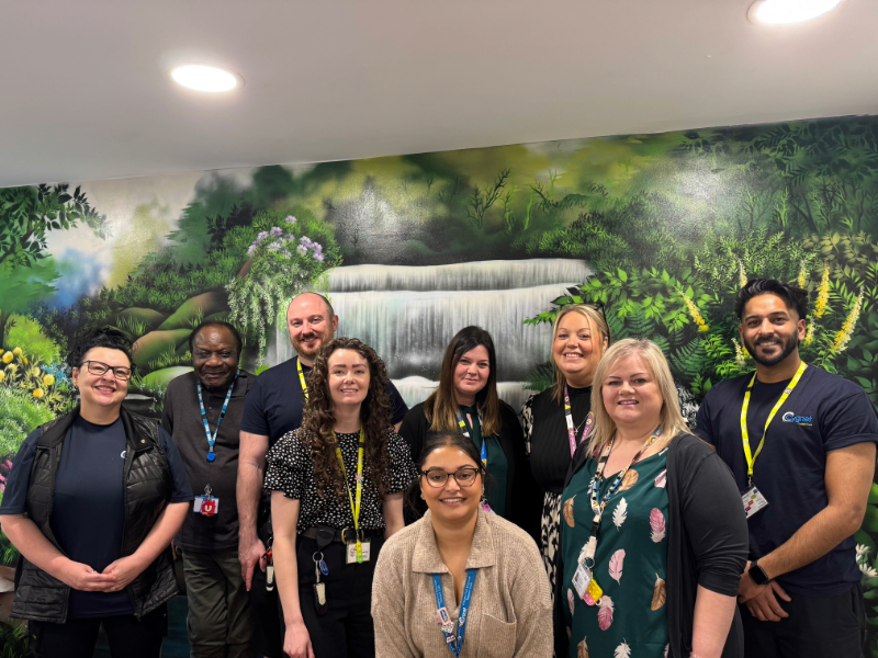 Cygnet Manor team smiling and celebrating the Triangle of Care award in front on colourful waterfall mural