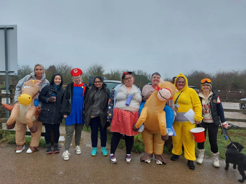 Group from Cygnet Maple House in colourful fancy‑dress costumes during a charity walk at Gedling Country Park, raising funds for The Friary; a red collection bucket and a small dog are visible.