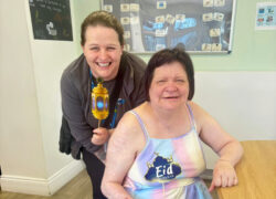 A member of the team and service user at a decorated table celebrating Eid, with one holding a colourful lantern and the other holding a sign saying 