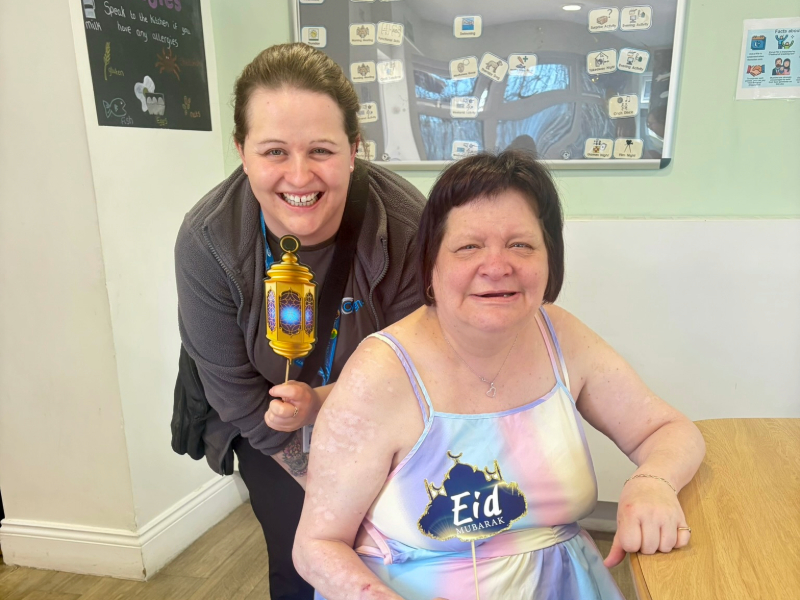 A member of the team and service user at a decorated table celebrating Eid, with one holding a colourful lantern and the other holding a sign saying 