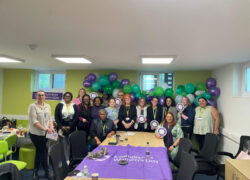 A group of Cygnet Women's Network colleagues standing together in a meeting room decorated with green and purple balloons for an International Women’s Day celebration