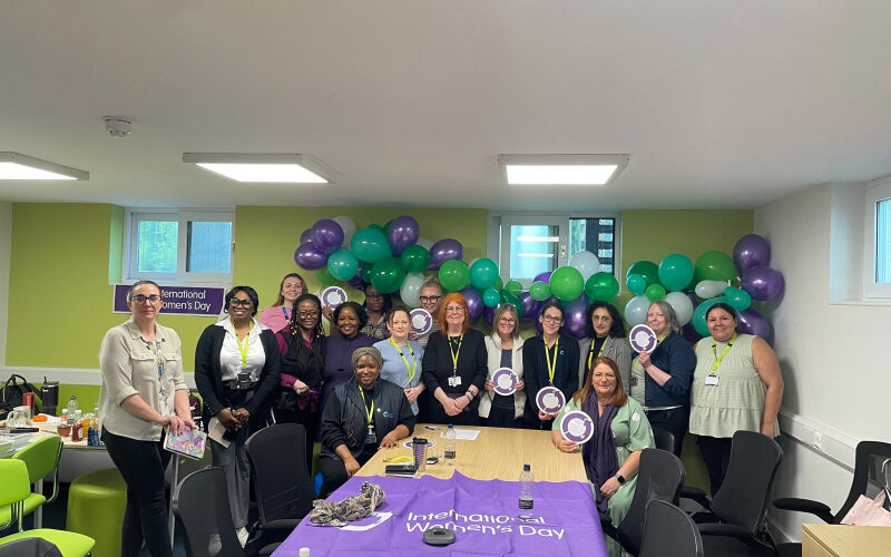 A group of Cygnet Women's Network colleagues standing together in a meeting room decorated with green and purple balloons for an International Women’s Day celebration