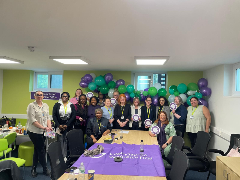 A group of Cygnet Women's Network colleagues standing together in a meeting room decorated with green and purple balloons for an International Women’s Day celebration