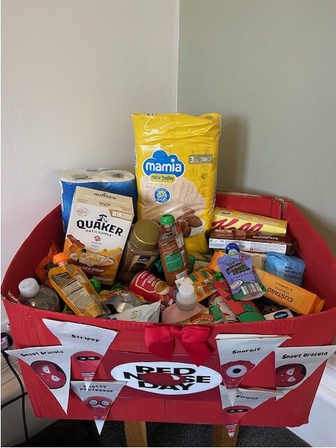 A handmade Red Nose Day donation box filled with food and household items, including cereal, nappies, toiletries and packaged goods, created by service users at Gledholt to donate to a local food bank.