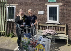Two staff members from Gledholt standing outside The Welcome Centre in Huddersfield next to a trolley filled with bags of donated food and supplies.