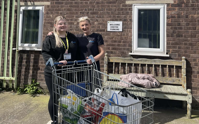 Two staff members from Gledholt standing outside The Welcome Centre in Huddersfield next to a trolley filled with bags of donated food and supplies.