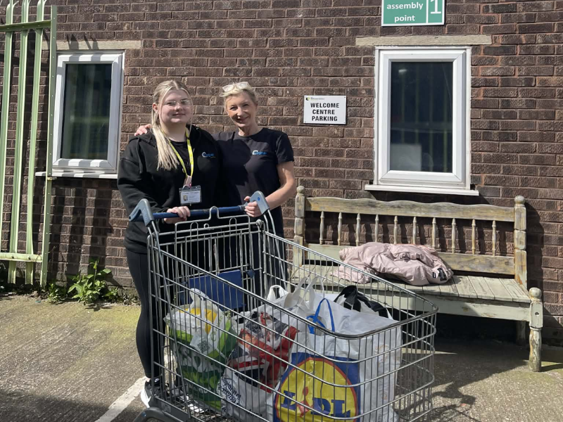 Two staff members from Gledholt standing outside The Welcome Centre in Huddersfield next to a trolley filled with bags of donated food and supplies.