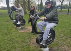 Two individuals supported at Toller Road smiling while on workout equipment at the outdoor gym with Support Worker Grace stood between them.