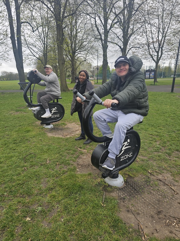 Two individuals supported at Toller Road smiling while on workout equipment at the outdoor gym with Support Worker Grace stood between them.