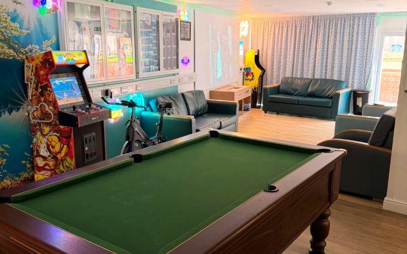 The social hub at Cygnet Lodge Lewisham from the doorway, including pool table, seating areas, arcade machine and exercise bike in soft lighting