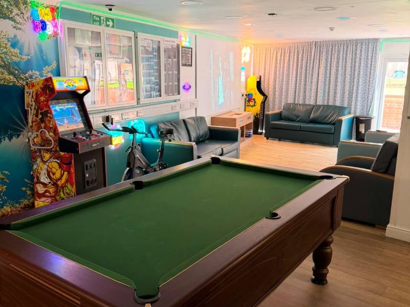 The social hub at Cygnet Lodge Lewisham from the doorway, including pool table, seating areas, laser projector, arcade machine and exercise bike in soft lighting