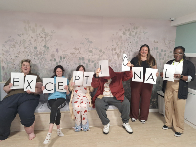 Six service users at Cygnet Wallace Hospital hold individual letter cards spelling "EXCEPTIONAL" as they celebrate the hospital's Healthcare Improvement Scotland inspection result.